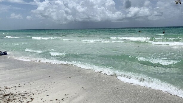 Ein Strand mit klarem türkisblauem Wasser, einem sandigen Ufer und einem teils bewölkten Himmel.