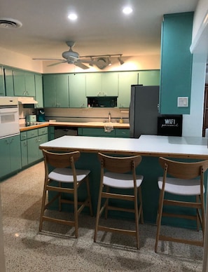 A kitchen with teal cabinets, a white countertop, and a black refrigerator. There are three wooden chairs with white cushions.