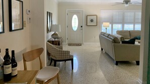 Ein Wohnzimmer mit einer Couch, einem Sessel, einem Couchtisch und einem Essbereich mit Tisch und Stühlen. Eine Tür mit rundem Fenster, ein Deckenventilator und gerahmte Bilder sind an den Wänden zu sehen.