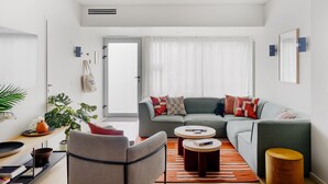 Een moderne woonkamer met een grijze bank, twee fauteuils, een bijzettafeltje en een klein houten krukje. Links staat een plant en op de achtergrond een raam met dunne gordijnen.