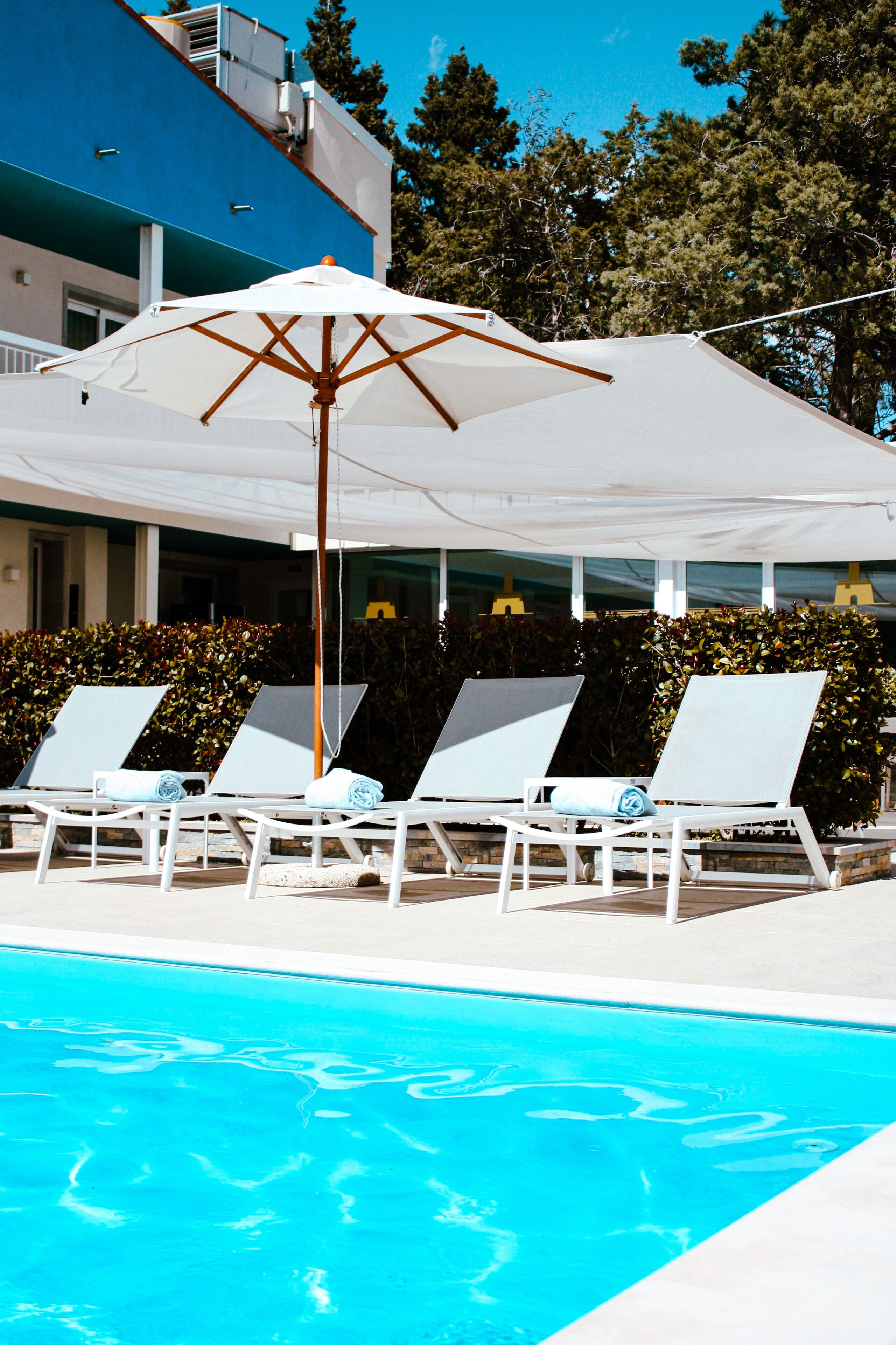 En swimmingpool med solstolar och parasoller, omgiven av en byggnad och träd.