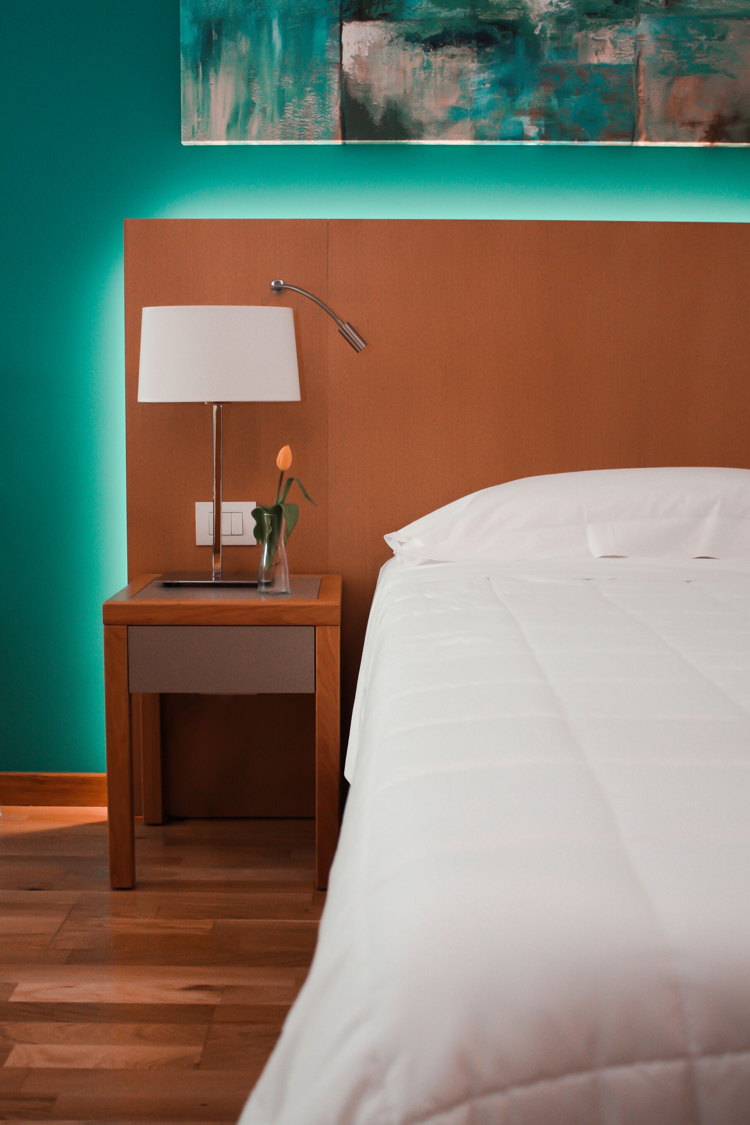 A neatly made bed with a white comforter and pillows, a wooden nightstand with a lamp and a small vase with a flower, and a teal wall with a painting above the bed.