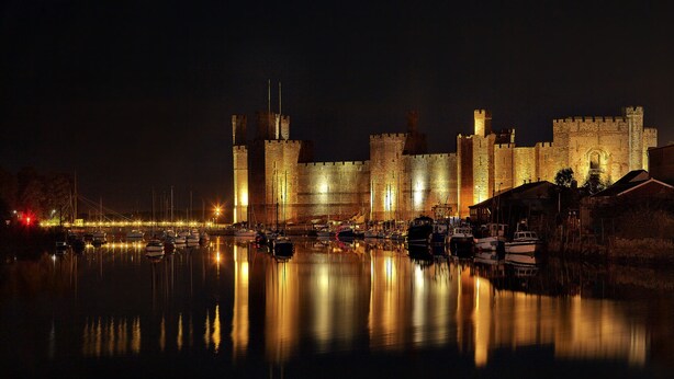 Um castelo histórico com fosso e barcos, iluminado à noite.