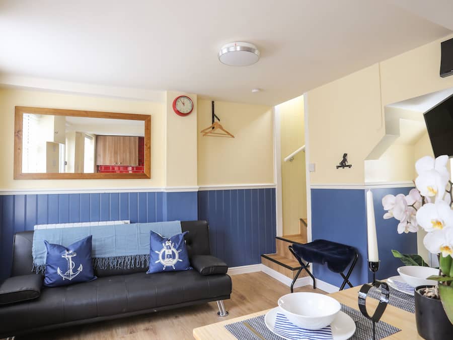 Ein Wohnzimmer mit einer schwarzen Couch, blauen Kissen, einem Holztisch, einem Esstisch mit weißer Schüssel und Tellern, einer Pendelleuchte und einem Spiegel an der Wand.