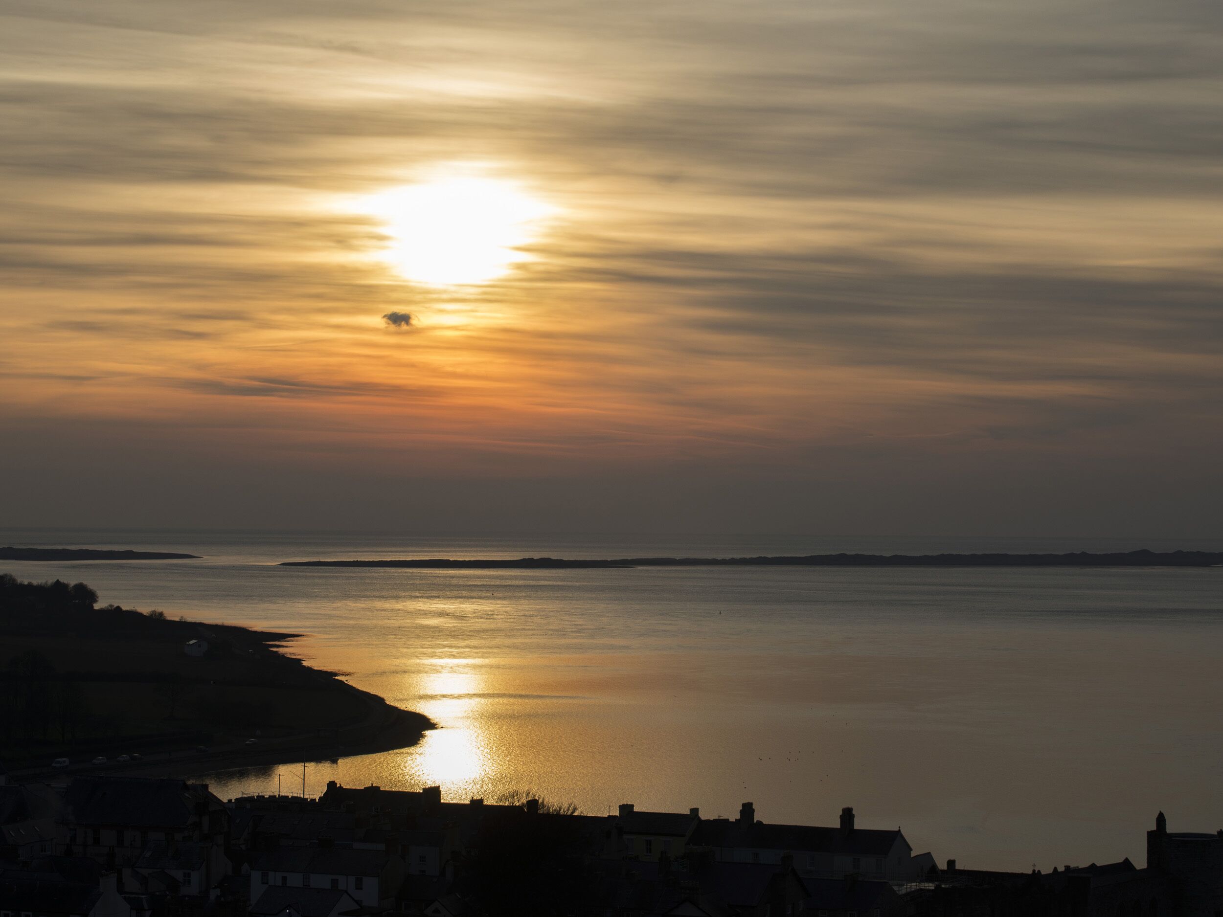 A coastal sunset view with a calm sea reflecting the sun, a distant shoreline, and a town silhouette.