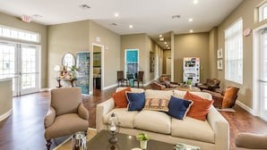 Amplia sala de estar con sofá color beige, sillones de cuero y una mesa de centro. Varias puertas conducen a otras habitaciones, y se observa una máquina expendedora.