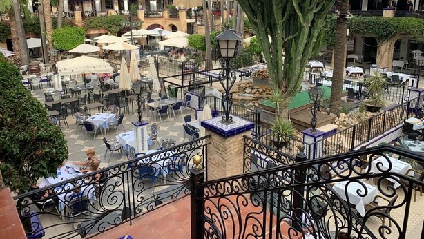 Un espace café en plein air, avec des tables et des chaises, entouré de palmiers et d’une balustrade en fer forgé.