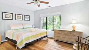 Un dormitorio con cama, piso de madera, cómoda, silla, ventilador de techo y ventana con vista a árboles.