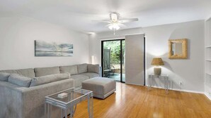 Un moderno salón con un sofá seccional gris, una mesa de centro de vidrio, un ventilador de techo y vistas al exterior a través de un ventanal.