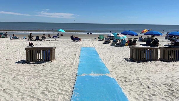 Una playa con un camastro azul que lleva al agua, sillas de playa, sombrillas y estructuras de madera.