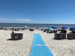 Ein Strand mit einer blauen Liegefläche, die zum Wasser führt, Liegestühlen, Sonnenschirmen und Holzkonstruktionen.
