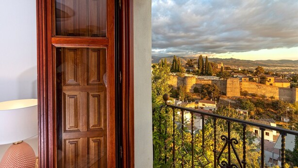 Una puerta de madera con un panel de vidrio, un balcón con baranda de hierro forjado y vistas a un pueblo histórico con edificios y árboles.