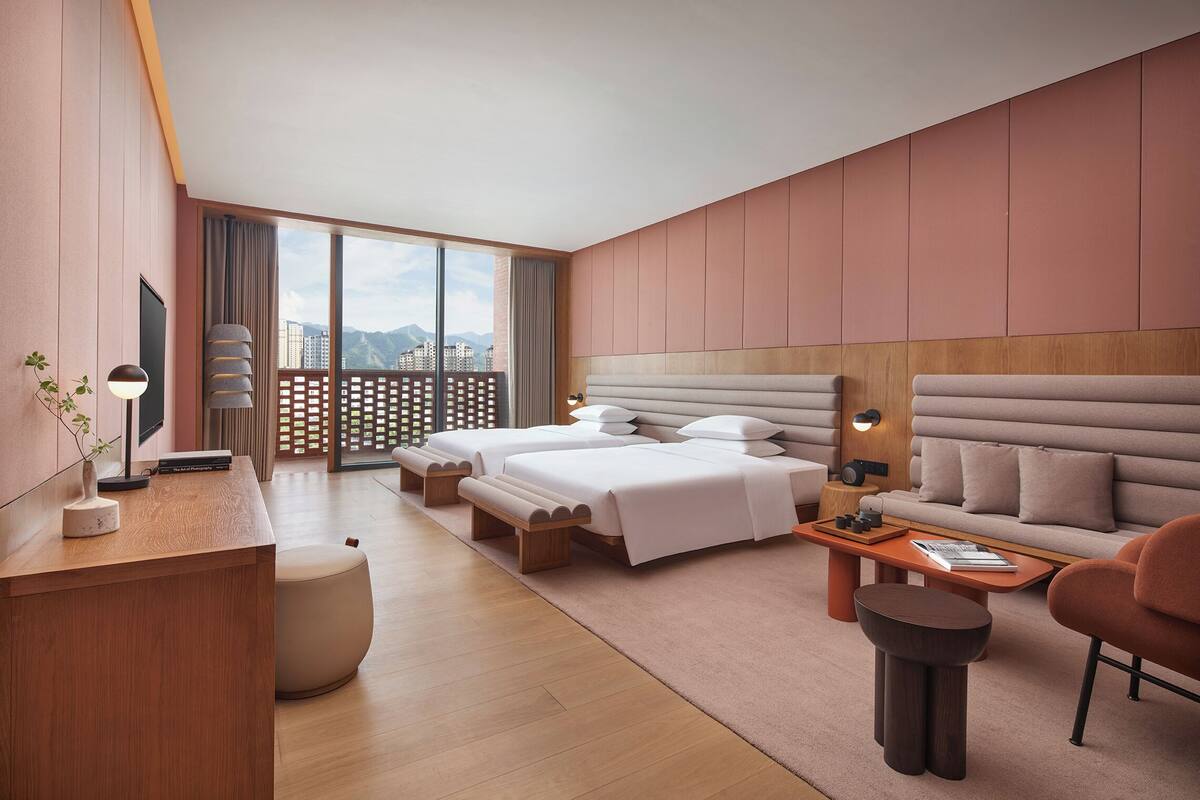 A modern hotel room with two beds, a sofa, a desk, and a view of the city.
