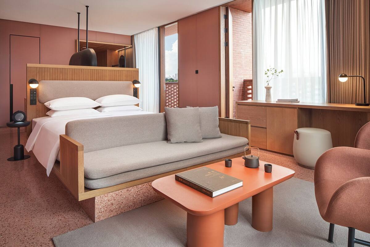 A modern hotel room with a bed, sofa, desk, and a view of the city.
