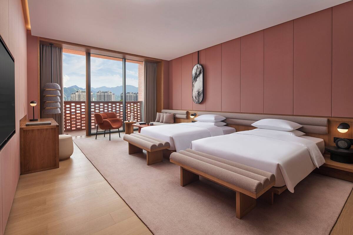 A modern hotel room with two beds, a desk, a chair, and a balcony with a city view.