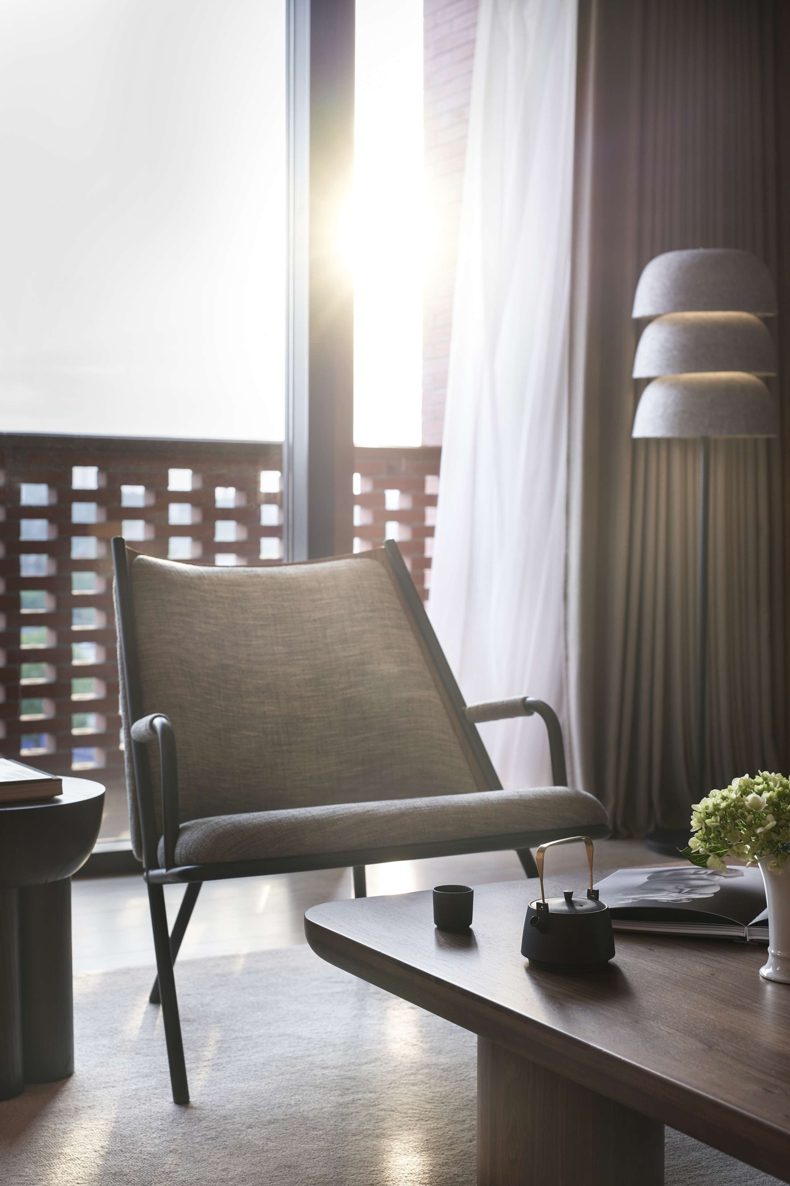 Una silla moderna con asiento y respaldo acolchados, una mesita con tetera y taza, y una lámpara con pantalla blanca.