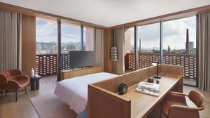 A modern hotel room with a large bed, a desk with a chair, a television, and large windows offering a city view.