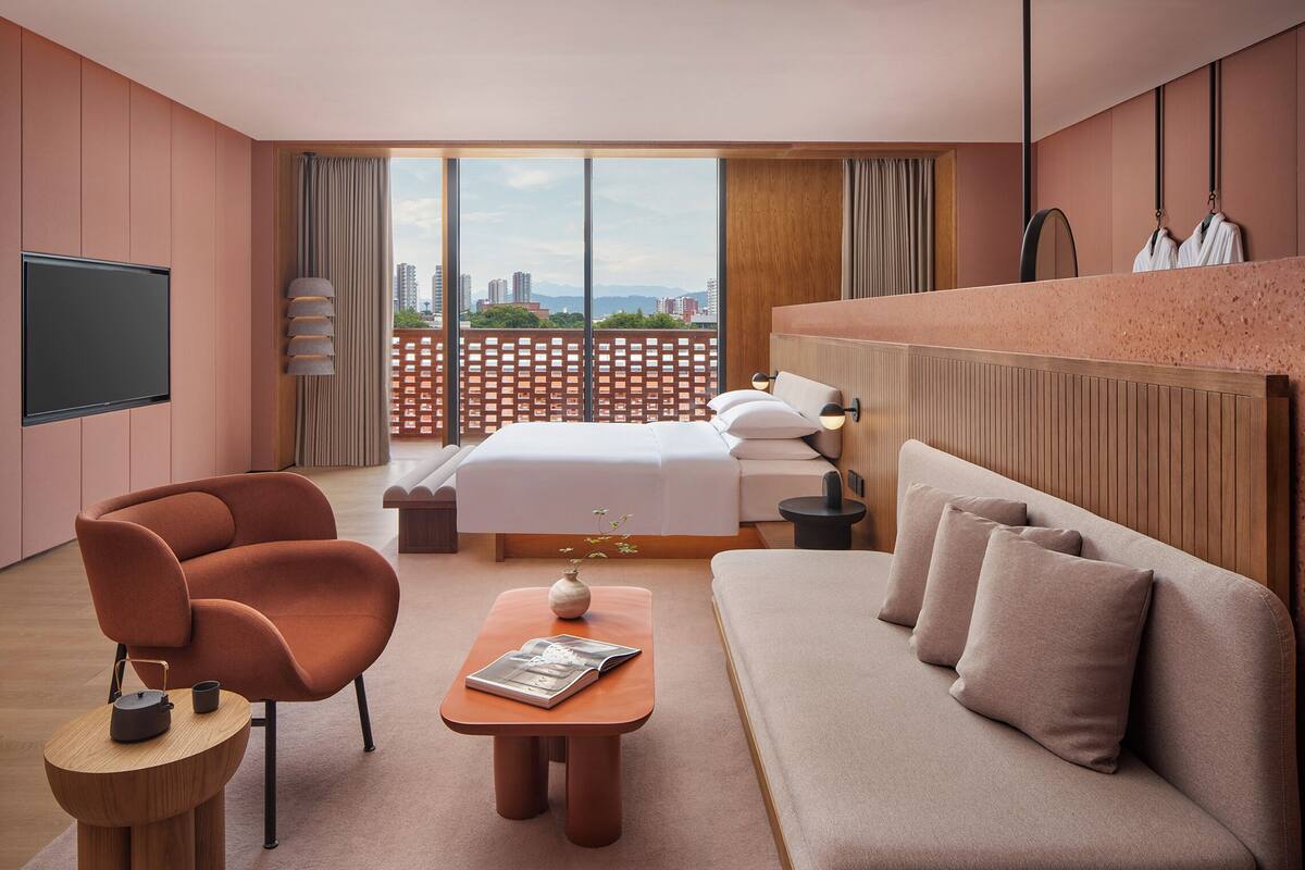 A modern hotel room with a bed, sofa, armchair, and a small table with a vase and magazine.