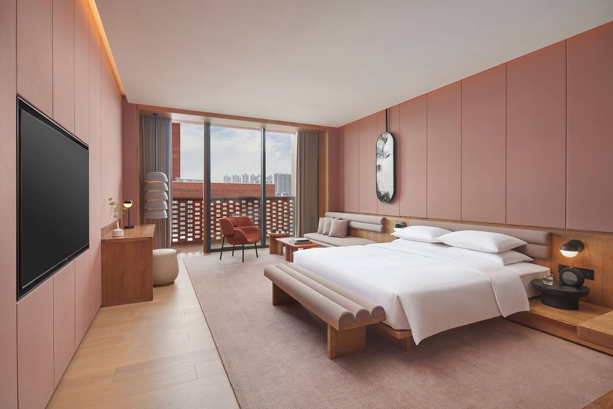 A modern bedroom with a large bed, a TV, a desk, and a balcony with a view of the city.