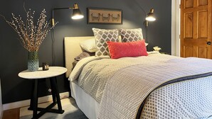 Un dormitorio con cama, mesita de noche, lámpara y un cuadro en la pared.