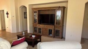 Un salon comprenant un grand meuble de rangement en bois servant de centre de divertissement, deux fauteuils blancs, une table basse avec des télécommandes et une bouteille d’eau.