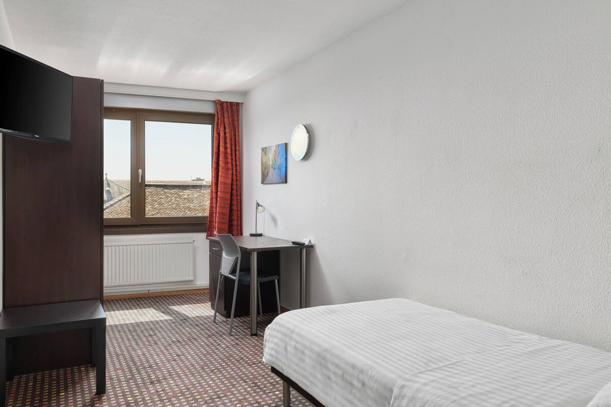 A hotel room with a bed, a desk, a chair, a TV, a window with curtains, and a radiator.