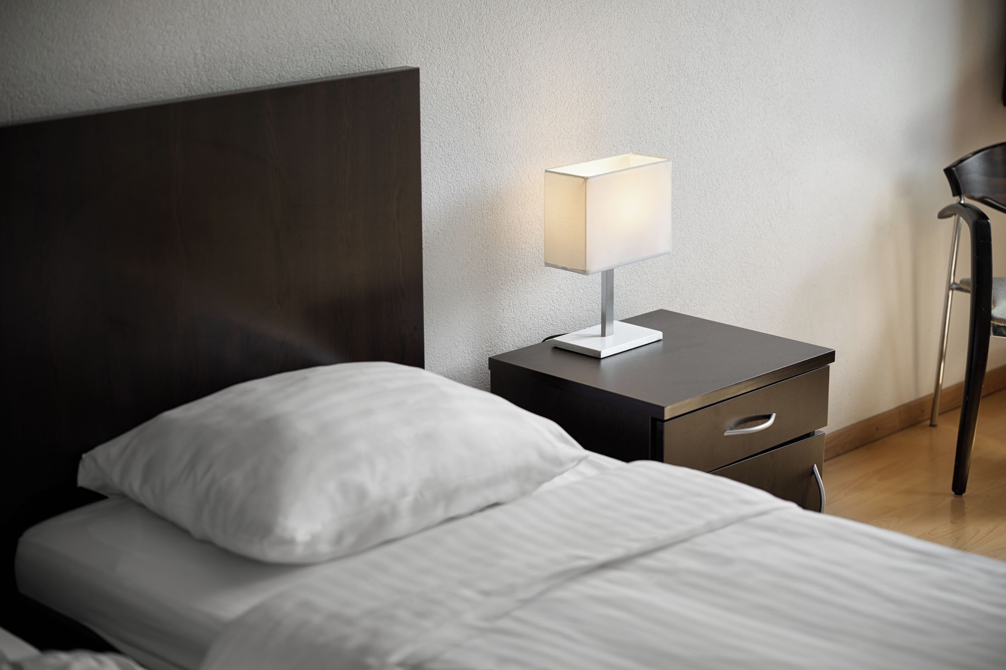 Ein ordentlich bezogenes Bett mit weißen Leinen, ein moderner Nachttisch mit Lampe und ein Sessel in einem Hotelzimmer.