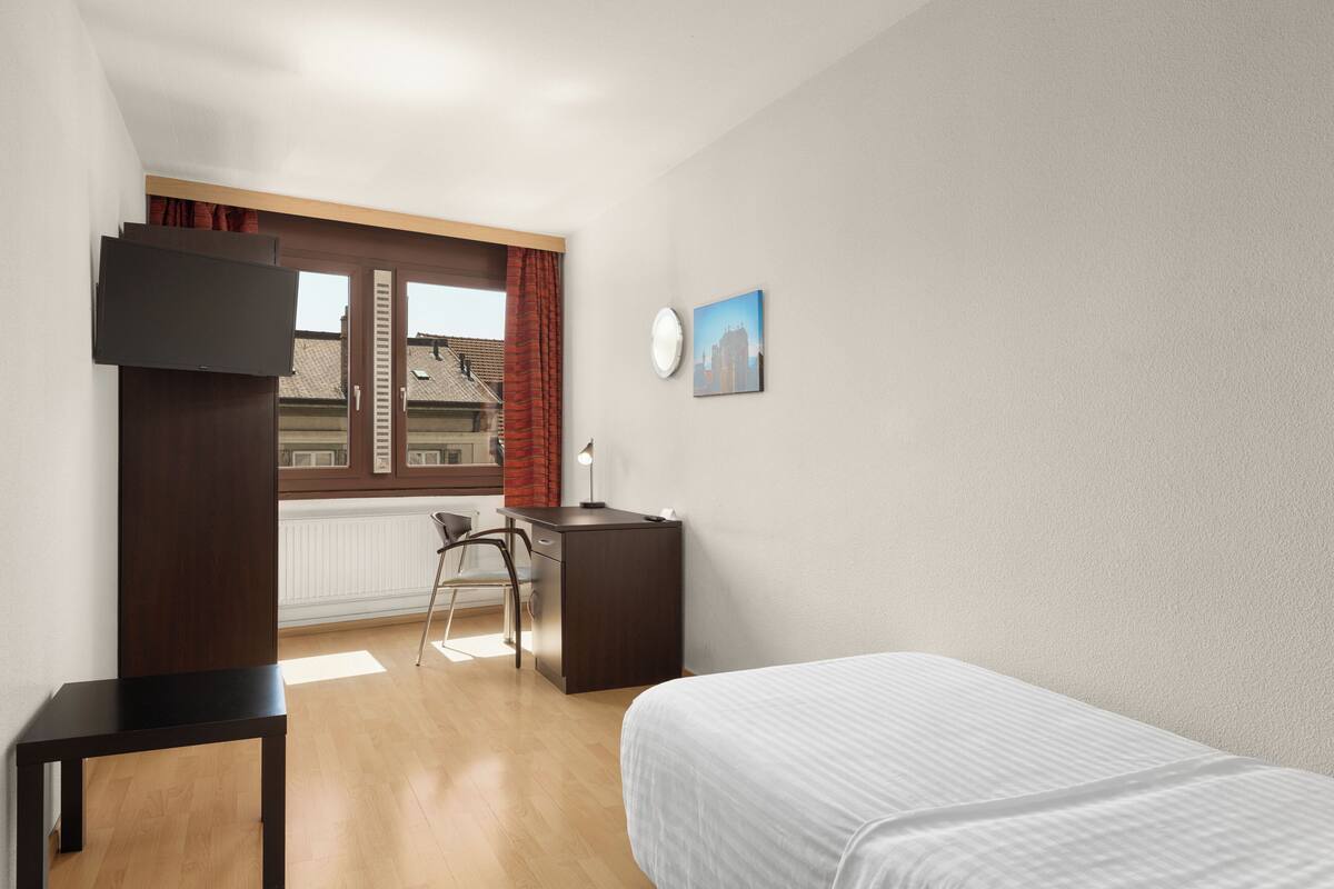 A bedroom with a bed, a desk, a chair, a TV, and a window with a view of buildings.