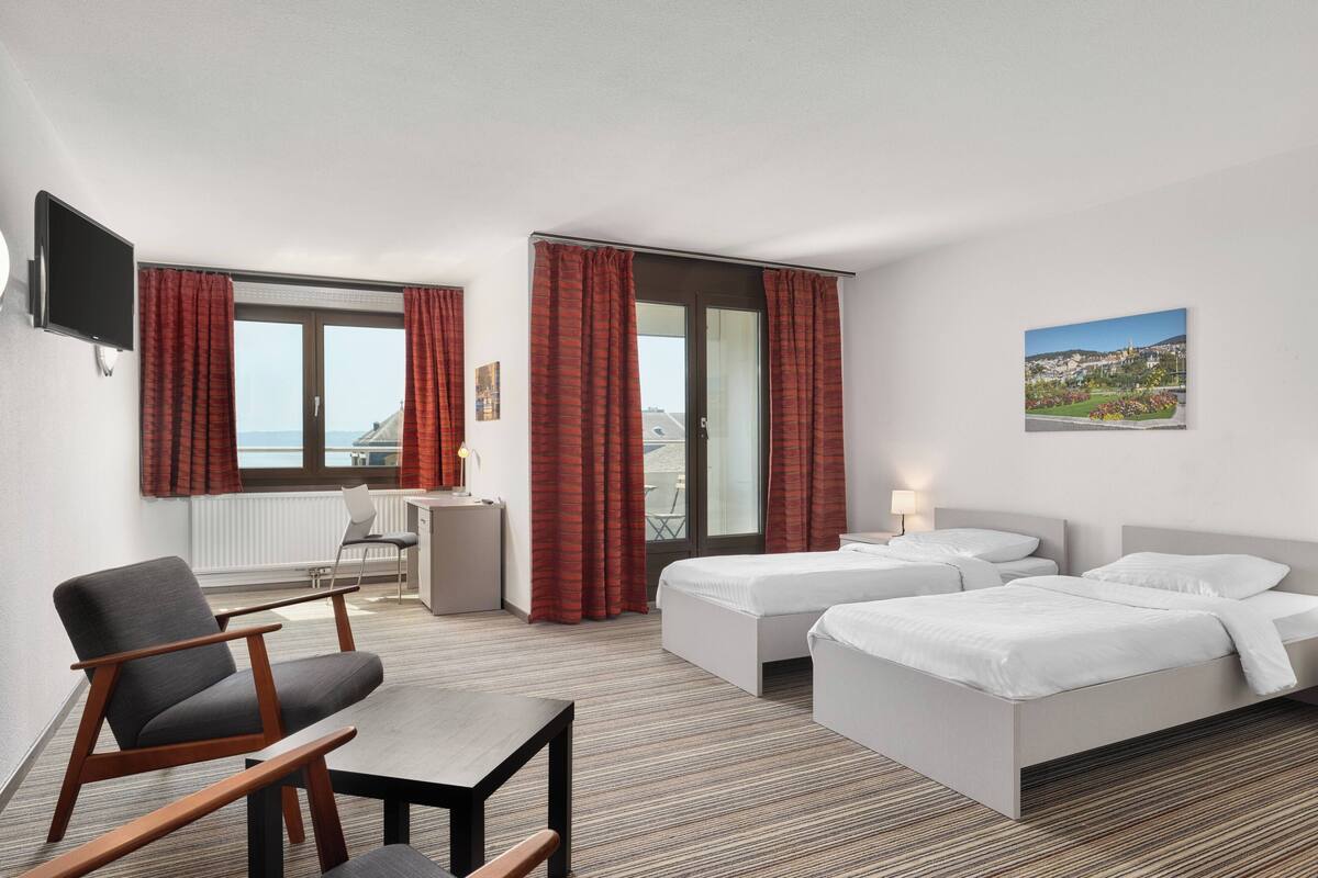 A hotel room with two beds, a desk, a chair, a TV, and a view of the city.