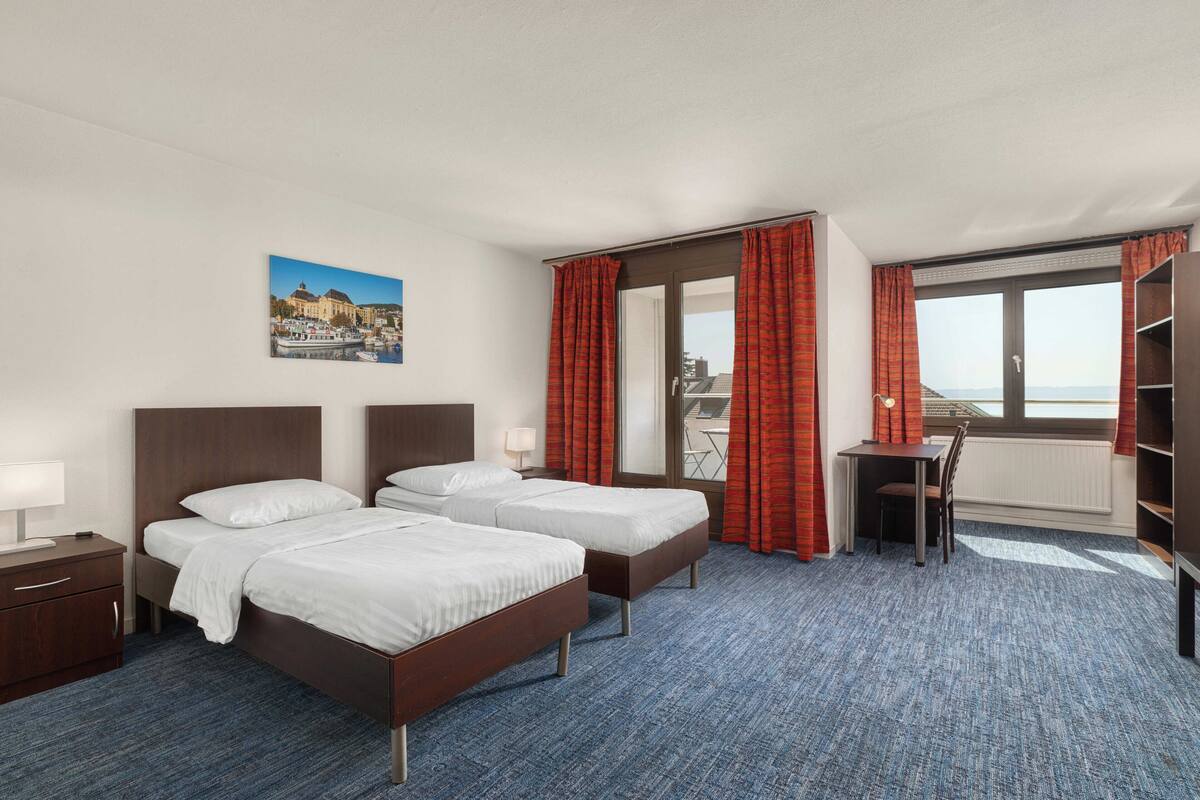 A hotel room with two beds, a desk, a chair, a window with curtains, and a painting on the wall.
