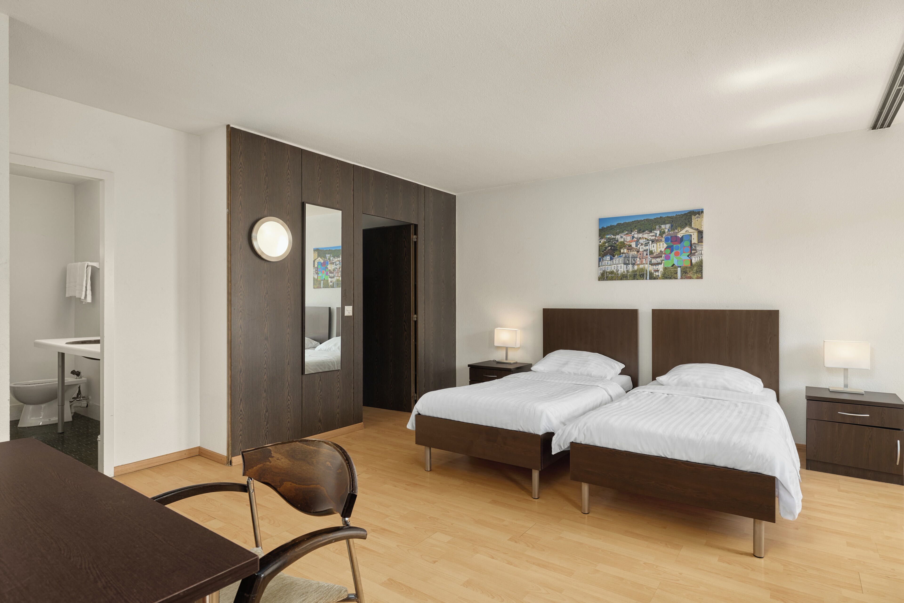 Habitación de hotel con piso de madera, dos camas, un escritorio, una silla, un baño con inodoro y lavamanos, y un cuadro en la pared.