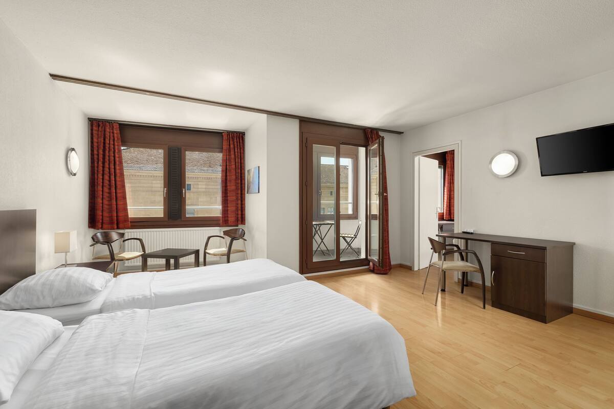 A hotel room with two beds, a desk, a chair, a TV, and a balcony with red curtains.