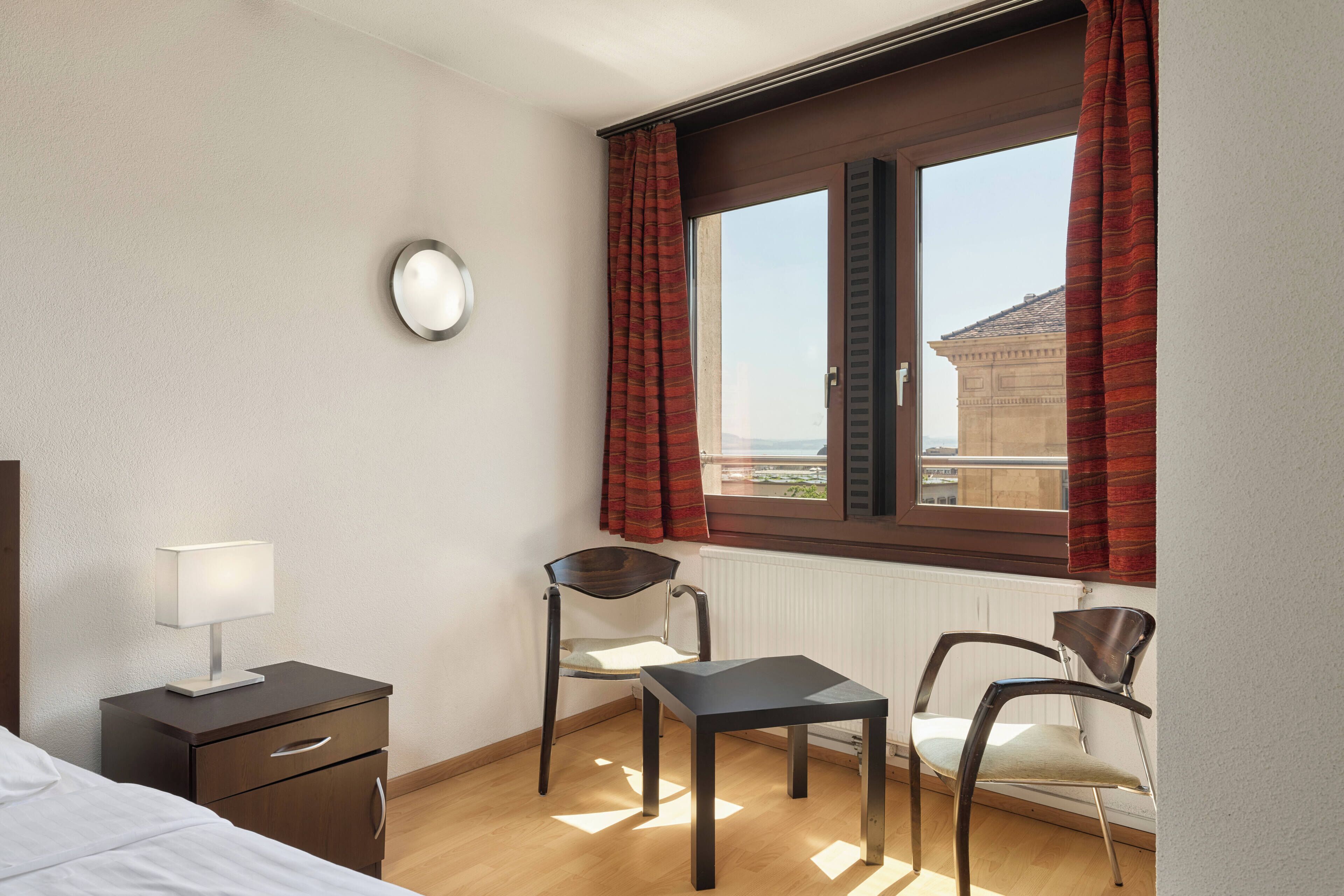 Una habitación de hotel con una cama, una mesita de noche con lámpara, dos sillas, una pequeña mesa, una ventana con cortinas y vistas a un edificio.