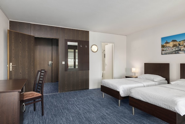 A hotel room with a desk, two beds, a chair, a wardrobe, a mirror, a clock, and a painting on the wall.