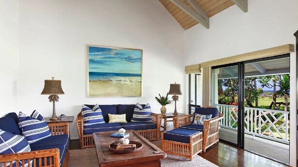 Amplia sala de estar con un gran ventanal que da a un jardín tropical, un cuadro con vista a la playa y techo de madera.