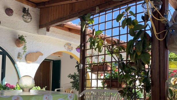 An outdoor patio with a wooden table covered by a floral tablecloth, a white plastic chair, and potted plants. - Cottage located along the coastal strip opposite Asinara. (Eden Beach)