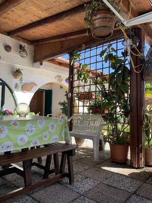 An outdoor patio with a wooden table covered by a floral tablecloth, a white plastic chair, and potted plants. - Cottage located along the coastline across from Asinara (Eden Beach)