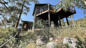 A two-story log cabin with a balcony, surrounded by trees and rocks. - NEW Entire Cabin, 🏔 & 🌊 Views + Hot Tub Downtown (Grand Lake)