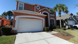 A two-story house with a red facade, a white garage door, and a small circular window on the second floor. - Terra Verde 116 (Kissimmee)