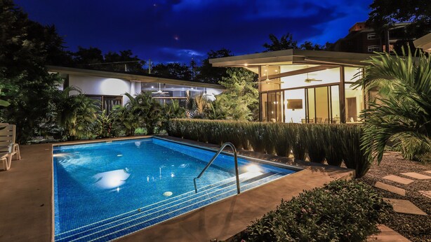 A swimming pool with a clear blue water, surrounded by a well-maintained garden and a modern building with large windows. - 55% OFF NOW Stylish 2BR Pool Retreat — Walk to Beach (Playa Langosta)