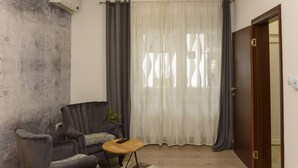 A living room with a grey armchair, a small round table, and a large window with sheer curtains.