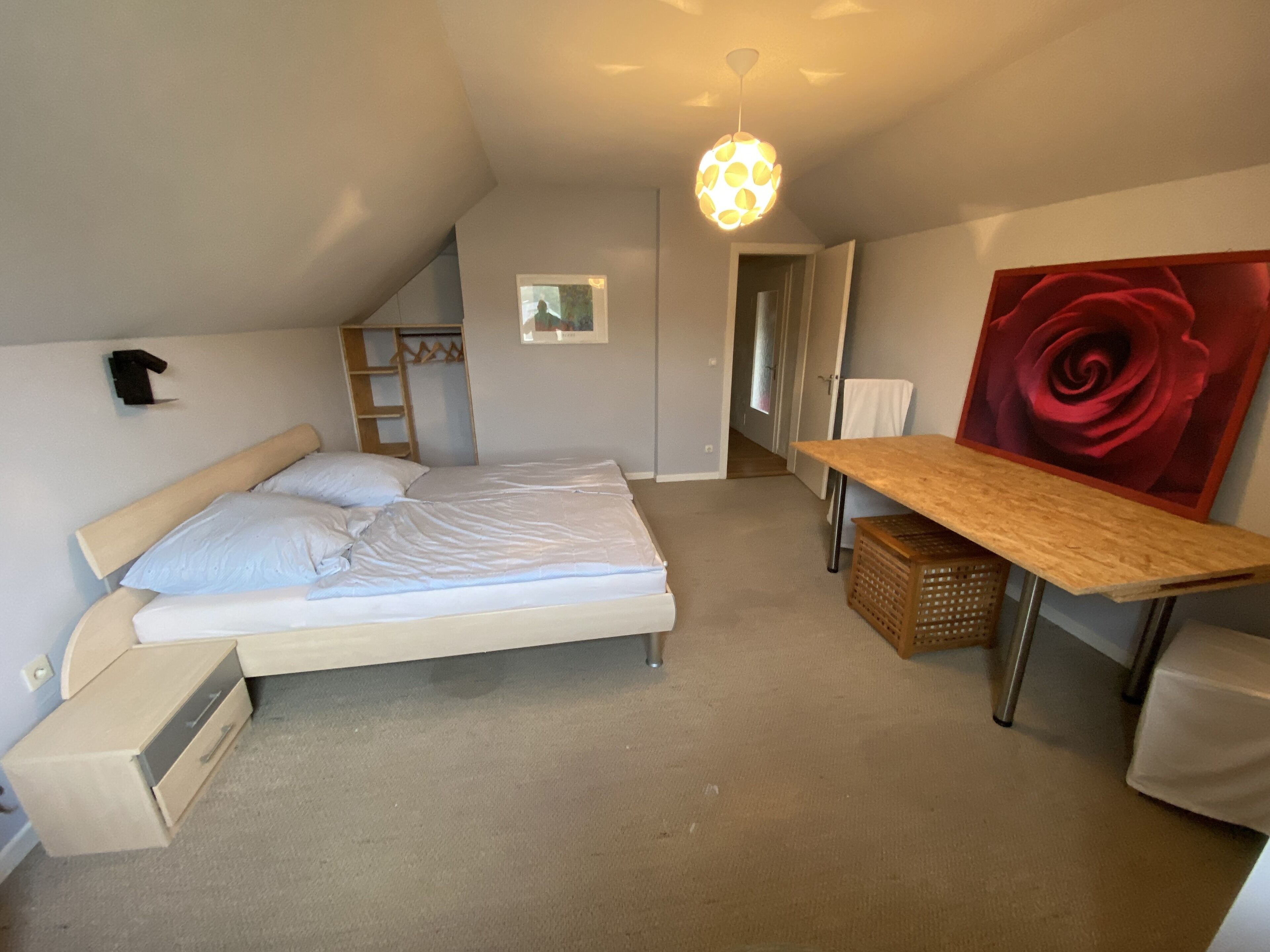 A bedroom with a bed, a wooden table, a red rose painting, and a ceiling light.