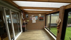 A covered patio area with wooden flooring, glass sliding doors, and a brick wall. - Cozy house with garden and terrace (Espelkamp)