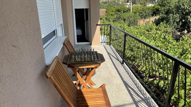 A balcony with a chess table and two wooden chairs, a closed window, and a view of greenery and buildings.