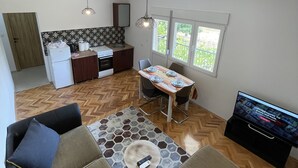 A modern living room with a sofa, a coffee table, and a TV. There is a dining area with a table and chairs. The room has wooden flooring and a kitchen in the background.