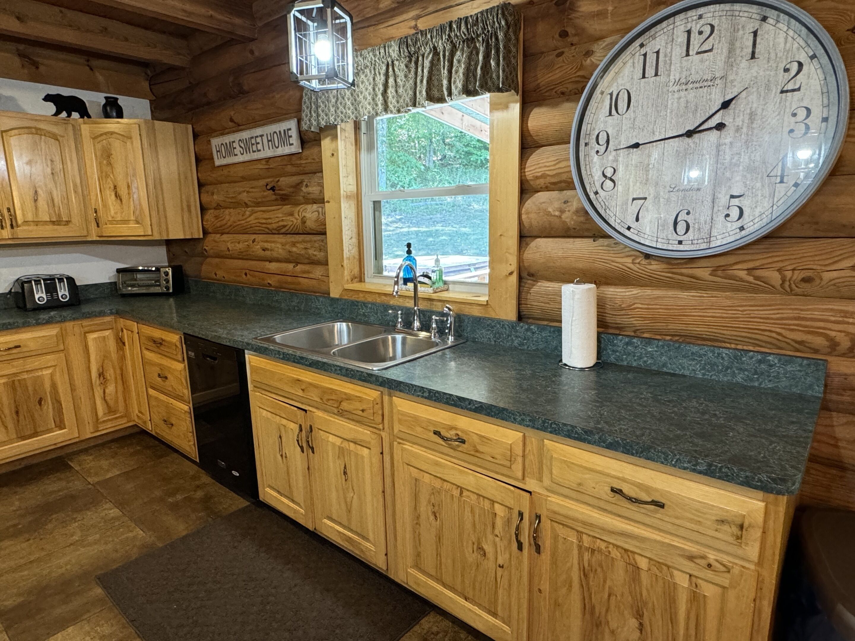 Une cuisine rustique aménagée dans une cabane en bois, avec une grande horloge, des placards en bois, un évier et une fenêtre donnant sur un paysage agréable.