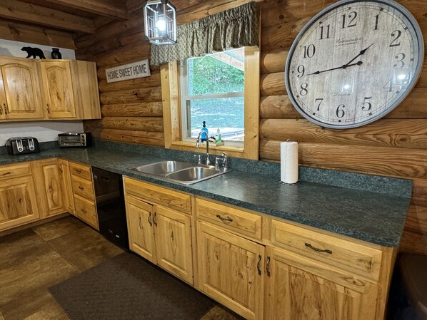 A wooden cabin kitchen with a large clock, wooden cabinets, a sink, and a window with a view.