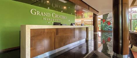 A reception desk at the Grand Crucero Iguazu Hotel with a green wall and a view of the hotel lobby.