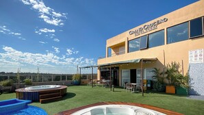 A hotel with outdoor hot tubs and a pool, surrounded by greenery and a clear sky.