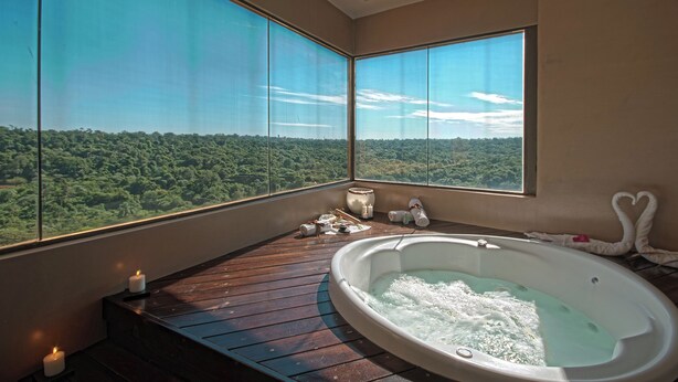 A room with a large bathtub, wooden deck, and a view of a forest through large windows.